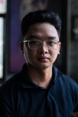 Obraz premium Portrait of a young Asian man wearing glasses, captured in a cozy indoor setting during daytime, with soft natural light in the background