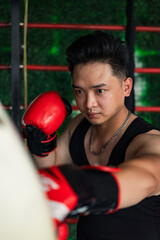 Young Asian man training in boxing with red gloves at a gym on a bright day while focusing on...
