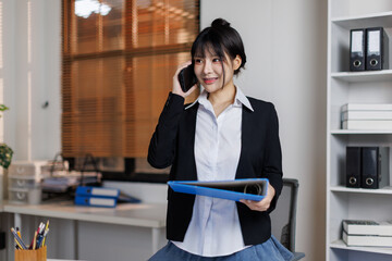 A cheerful, beautiful Young Asian businesswoman is talking on the phone with her client while...