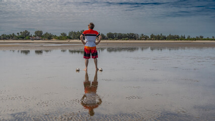 Low tide in the ocean. A man in shorts is standing in the shallow water, looking up. Flip-flops are...
