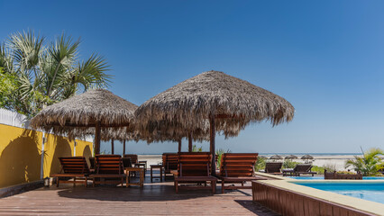 Sun beds under straw umbrellas by the swimming pool. A number of parasols on a sandy beach. The ocean is far away. Palm leaves against a clear blue sky. Copy  space. Madagascar. Morondava.