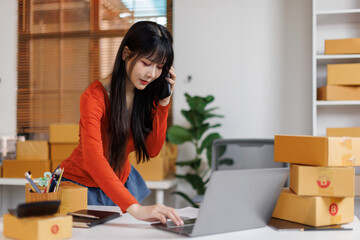 Cute Asian business owner talking on phone and taking order. Asian girl entrepreneur working at home office confirming the order on phone.
