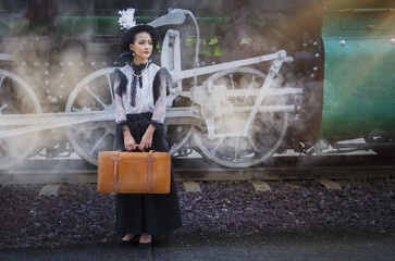 Young woman in vintage attire stands with suitcase beside steam train, evoking sense of nostalgia...