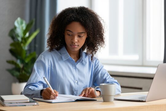 African american woman writing in notebook at desk home office studying online class