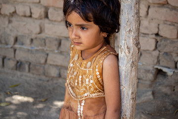 Portrait of a young rural girl standing near wooden post in front of brick wall, thoughtful...