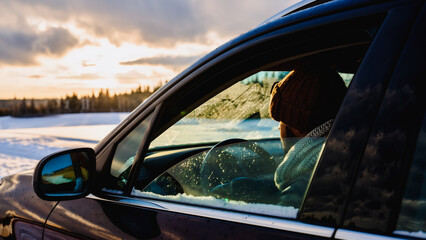 Snowy Sunset Reflection - Contemplative Driver's Moment, A person wearing a hat sits in the driver's seat of a car, gazing out the window during a snowy sunset.