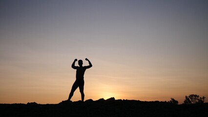 Silhouette of triumph: A lone figure stands silhouetted against a vibrant sunset, arms raised in a...