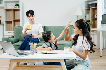 Asian parents cheering and guiding daughter through homework at home. Joyful teamwork, love, and...