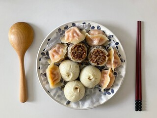 Overhead shot of assorted Chinese dim sum on a decorative plate, including dumplings, shumai, and steamed buns, served with chopsticks and a wooden spoon on a white table.
