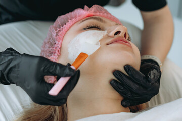 professional cosmetologist applies a creamy face mask on yiung woman face using a brush