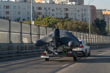 A white automobile towing a covered recreational boat on a trailer along a busy urban highway next...