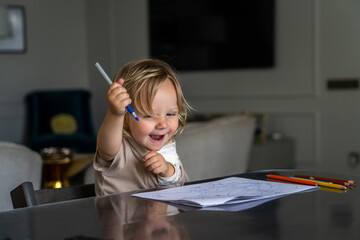 A joyful toddler engaging in creative expression, holding a blue pen above scribbled paper at a...