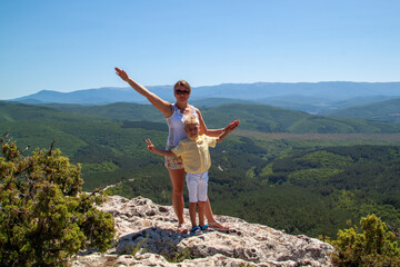 Joyful mother and son posing on a rocky mountain peak, overlooking a vast green forest landscape...