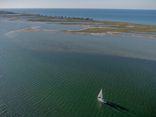 White sailboat glides across calm green-blue waters near the natural coastline of Graswarder in Heiligenhafen, Germany with sandbanks, tidal pool  and wide horizon.