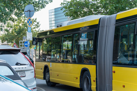 Germany Berlin July 19, 2025. A yellow articulated BVG bus drives past parked cars and German street signs in a busy urban setting. A mix of modern traffic and public transport is visible under green 
