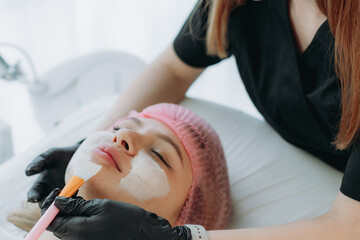 professional cosmetologist applies a creamy face mask on yiung woman face using a brush