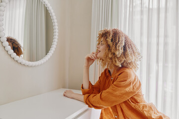 A thoughtful young woman with curly hair in an orange outfit gazes into a mirror, reflecting on her...