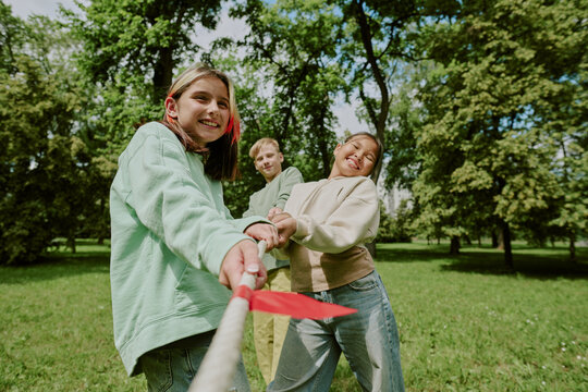 Group of multiethnic children playing tug of war in park, smiling and pulling rope together, enjoying outdoor activity with classmates, trees and grass visible in background