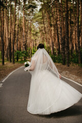 A bride in a white fluffy dress with a long veil and a bouquet of flowers walks along a long...