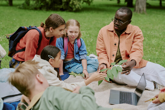 Black male young adult teacher sitting on grass in park engaging diverse group of children in outdoor learning activity, children examining plant leaves near open laptop computer