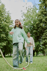 Two preteen girls, one Caucasian and one Asian, playing with jump rope outdoors in park, smiling...