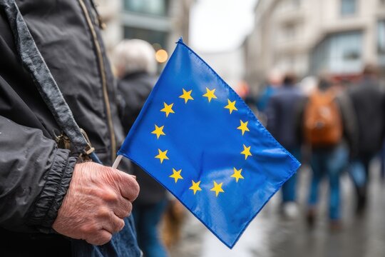 A man in a black jacket holds a small blue EU flag with yellow stars on a daytime street