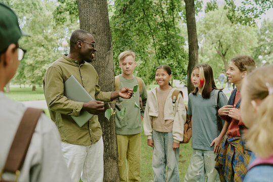 Middle aged Black man teacher holding laptop engaging with diverse group of children and teenagers standing in park listening attentively during outdoor educational activity