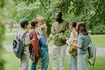 Group of diverse children and Black male teacher standing outdoors in park, teacher showing plant to students, children holding notebooks and magnifying glass, participating in educational activity