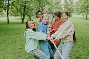 Group of diverse children smiling and playing tug of war together in park, pulling rope outdoors,...