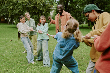 Group of multiethnic children playing tug of war outdoors with Black male teacher supervising, kids smiling and laughing while pulling rope together in green park setting