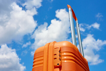 Bright orange suitcase with extended handle stands against blue sky with white clouds. Concept of...