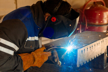 A welder in protective clothing and a mask performs manual arc welding of metal, sparks fly off,...