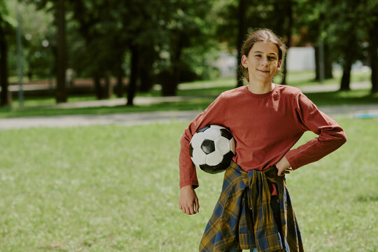 Portrait of Caucasian teenage boy standing outdoors in park holding soccer ball under arm, smiling at camera, wearing casual clothes, surrounded by green grass and trees