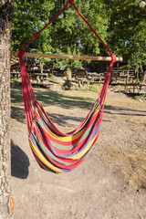 Colorful striped hanging hammock chair tied to tree in sunny forest park with picnic benches in background