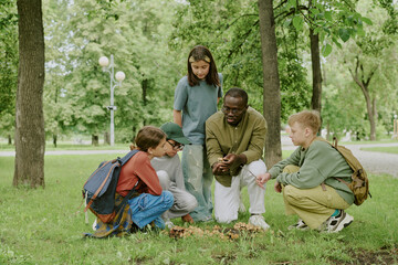 Black male young adult teacher kneeling on grass in park showing group of multiethnic children and...