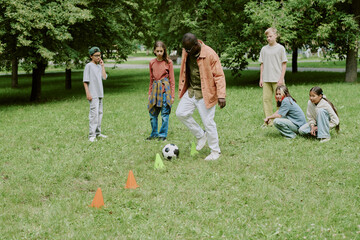Group of diverse children standing and sitting on grass in park, watching Black male teacher demonstrating soccer skills with ball and cones during outdoor activity
