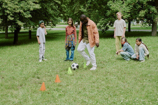 Group of diverse children standing and sitting on grass in park, watching Black male teacher demonstrating soccer skills with ball and cones during outdoor activity