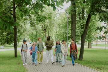 Group of diverse children walking with Black male teacher in park, kids talking and smiling while teacher engaging with students outdoors, trees and greenery surrounding pathway