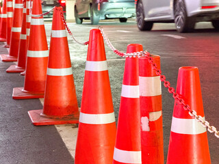 Row of orange traffic cones with reflective stripes and connecting chain near parked vehicles,...