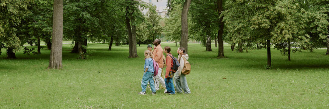 Group of multiethnic children walking with Black male teacher in park, kids carrying backpacks and listening attentively, outdoor educational activity in green natural setting - Powered by Adobe