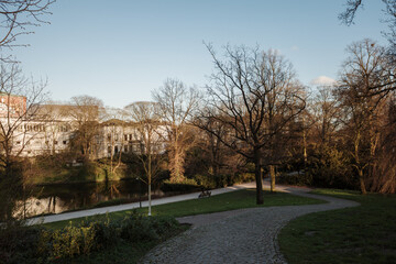 A scenic view of the Wallanlagen park in Bremen, Germany, during early spring or late autumn. A cobblestone path winds through the park next to the historic city moat (Stadtgraben). 
