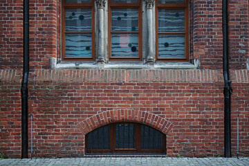 The detailed facade of an old, red brick building featuring ornate stone columns next to the upper windows and an arched basement window with bars.
