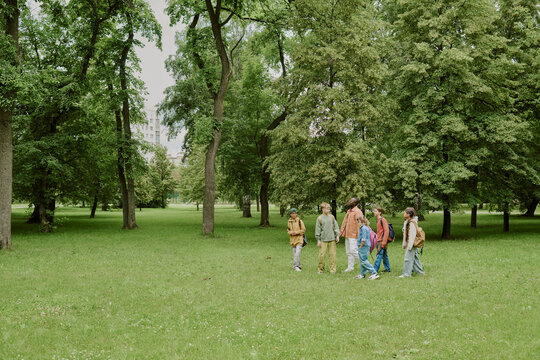 Group of multiethnic children and young Caucasian woman teacher walking together in green park, teacher leading children through grassy area surrounded by tall trees during outdoor activity