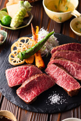 【黒毛和牛ステーキ】レアで美しく焼き上げた黒毛和牛モモ肉のステーキ。ジューシーな肉の旨みを引き立てるのは、素揚げした季節の野菜と彩り豊かな薬味。わさびや塩、日本酒とともに、和の美味を贅沢に味わう一皿です。