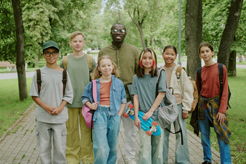 Diverse group of children and Black male teacher standing together in park smiling at camera,...