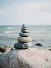 Fototapeta premium A carefully balanced stack of grey and brown stones sits atop a larger, pinkish rock near a calm sea under a pale sky