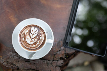 Beautifully crafted latte art sits in a white cup on rustic wooden table. Tablet rests, blending technology with enjoyment of coffee in serene, nature filled outdoor space.