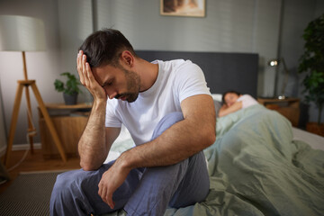 A man sits at the edge of the bed, distressed and thoughtful, while a woman lies under the covers,...