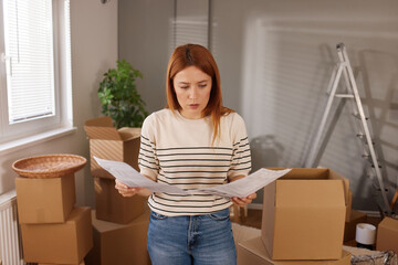A young woman stands in a partially unpacked room, holding important papers and looking concerned....