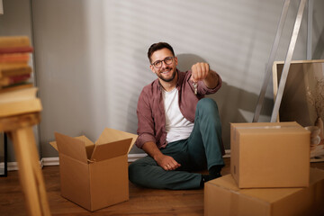 A joyful person sits on the floor in a newly moved-in home, surrounded by cardboard boxes, holding...